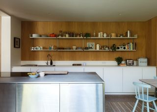 A stainless steel island in a modern kitchen with white lower cabinets and open shelving. On the shelves are lots of household items.