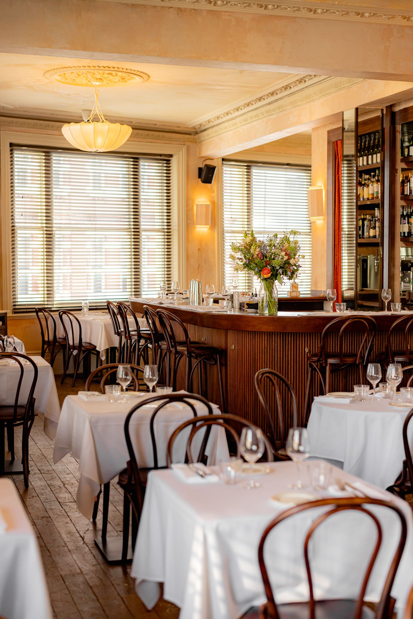 The charming-cool interiors of a French-style modern bistro with distressed plaster walls, lived-in wooden floors, elegant tables and chairs, colorful artworks, beige and red curtains, and sunset-hued sconces and pendant lights.