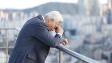 Concept image showing stressed male CEO standing with head in hands while leaning on a balcony railing overlooking a city skyline.