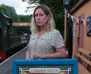 Vivienne (Anna Crilly) stands on the platform of the train station next to a vintage carriage, leaning on a sandwich board that is advertising the upcoming murder mystery production
