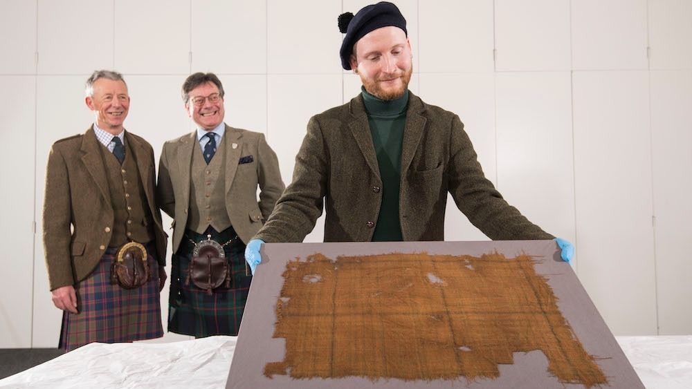 Peter MacDonald, head of research and collections and John McLeish, chair of The Scottish Tartans Authority and James Wylie, an assistant curator at V&amp;A Dundee, examine the faded tartan.