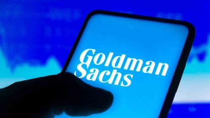 A person looking at a logo of Goldman Sachs on their phone, with a stock chart as the background