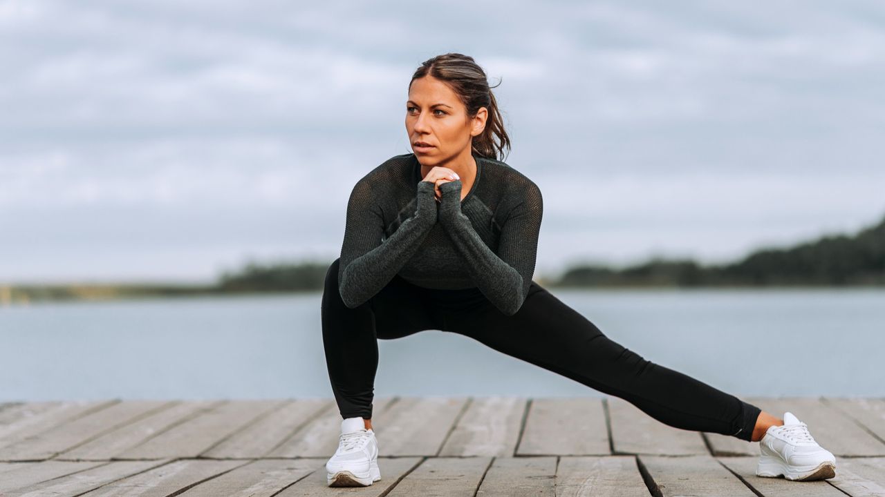 Woman wearing black leggings and a grey shirt performs a side lunge. Her hair is in a ponytail and her trainers are white.