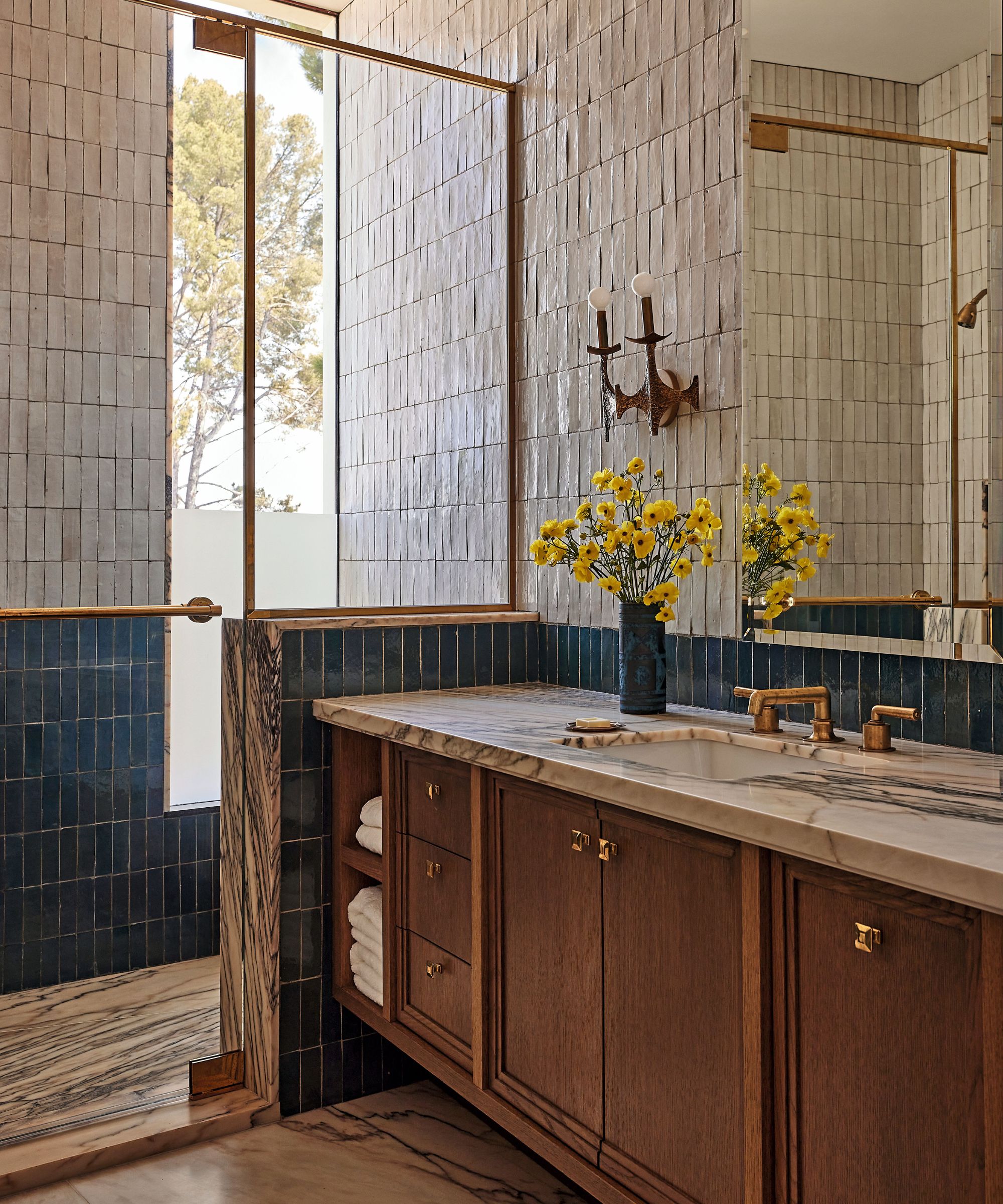 Bathroom vanity with marble countertop, rich wood cabinetry, brass hardware, and a textured wall of off-white and deep teal zellige tiles