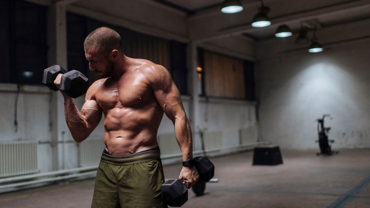 A man performing bicep curls with a pair of dumbbells with no top on