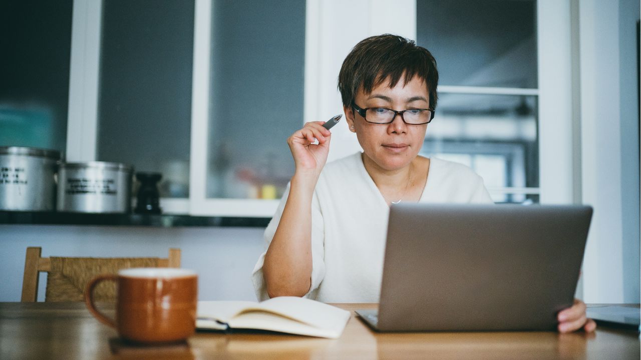 An older woman looks at her laptop while sitting at her kitchen table.