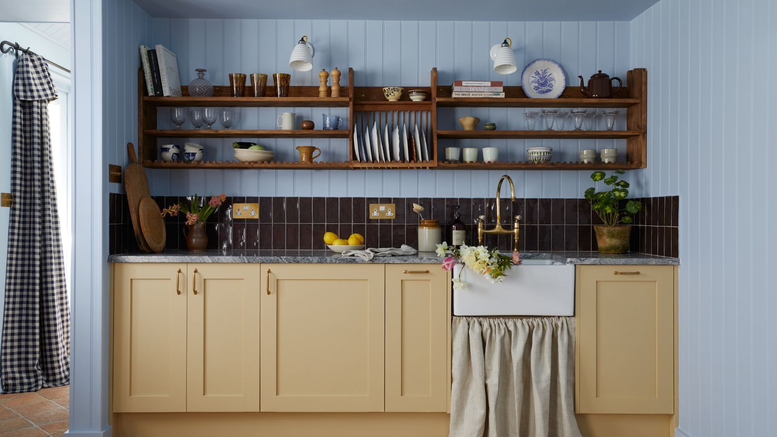 A cozy cottage kitchen with powder blue color-drenched panelled walls and butter yellow kitchen cabinets. Open wood shelving and dark burgundy backsplash tiles. 