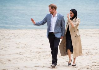 Meghan Markle wearing a tan trench coat and black dress and Prince Harry in black pants and a blue suit jacket walking on the beach and waving while holding hands