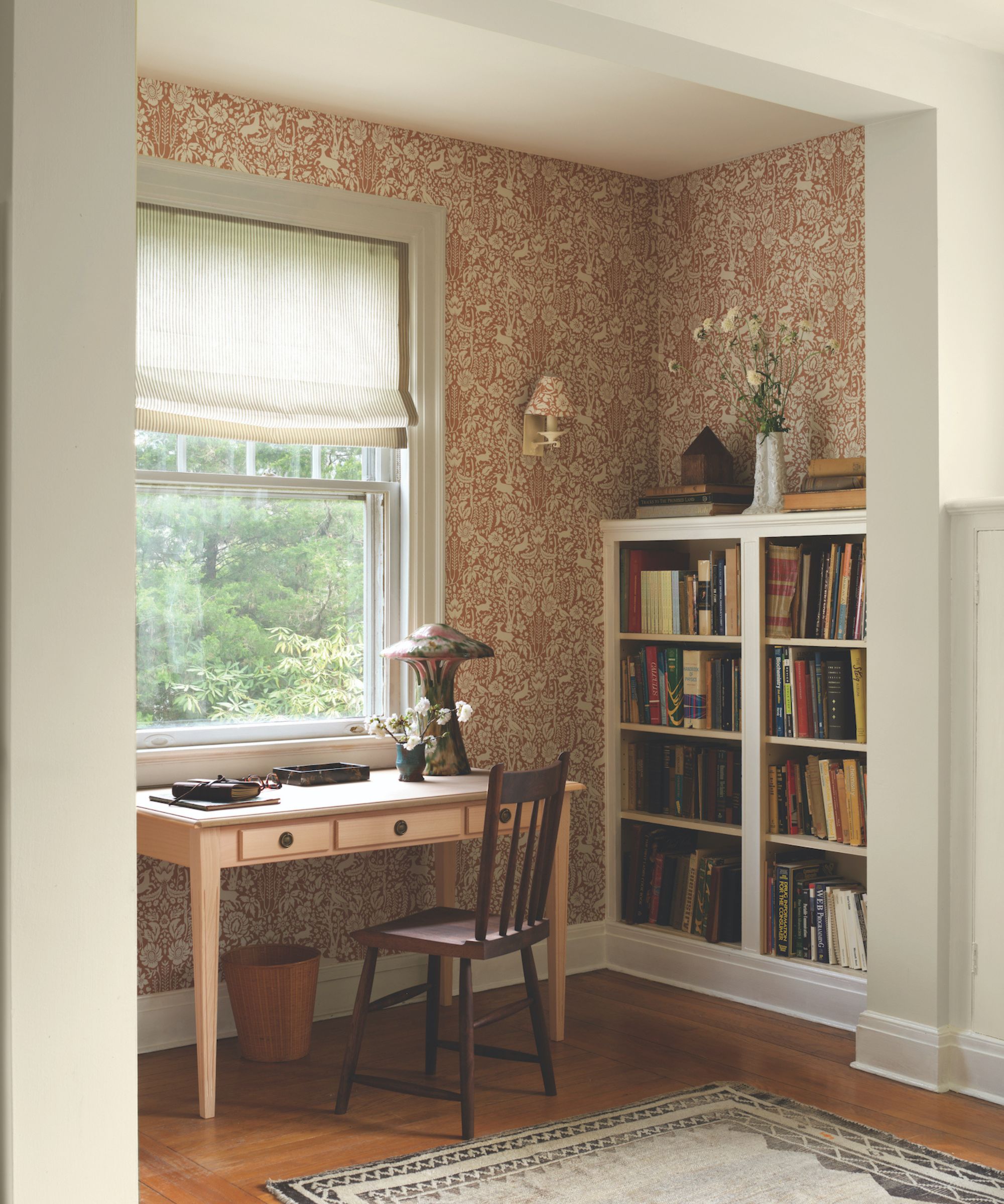 Home office with wood desk and chair, wood floor with woven rug and burgundy floral wallpaper