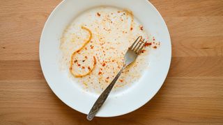 white plate sitting on a wooden table has remnants of red sauce and a single pasta noodle on its surface, alongside a fork