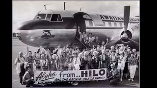 black and white image of old propeller commercial airline on a tarmac behind a large group of waiving people holding a sign that says 'aloha from HILO and the orchid isle of hawaii'