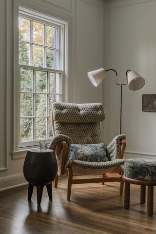 armchair with verdure print pillow and matching upholstered ottoman, with sculptural floor lamp and side table in front of a window