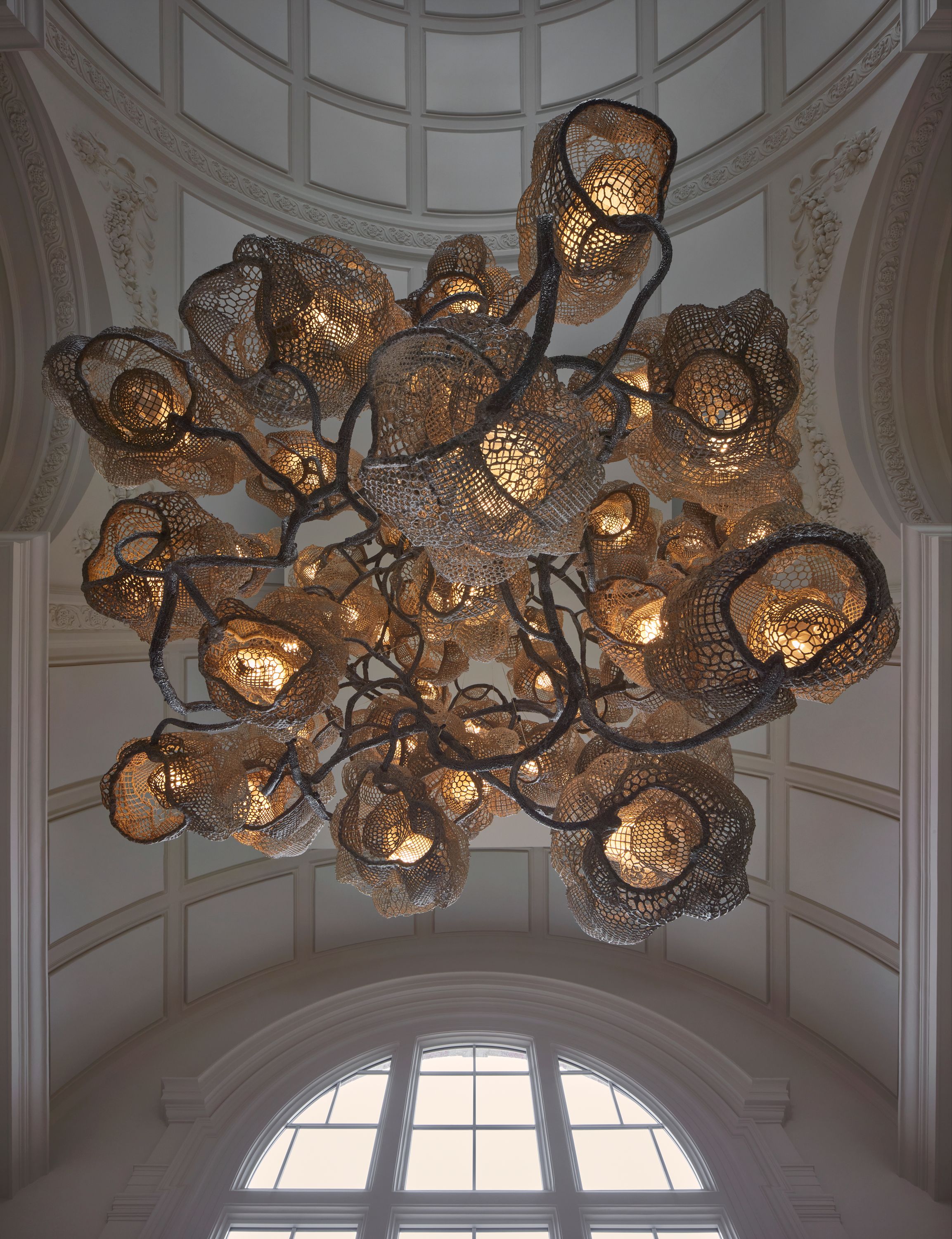 A sculptural chandelier made of multiple perforated, tree-like structures hangs in a vaulted hall and is captured from below.