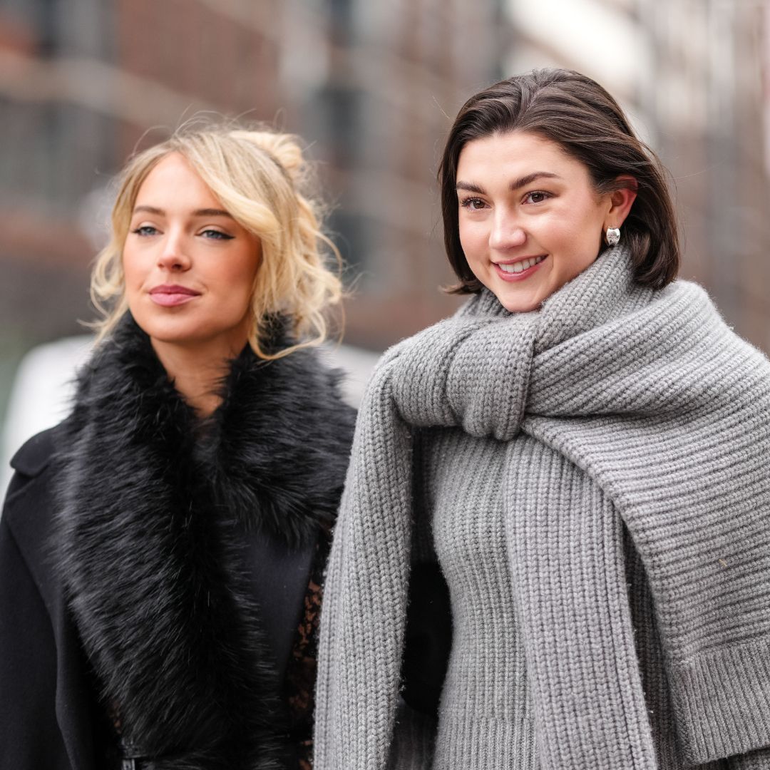 A guest wears black oversized Michael Kors coat, black faux fur scarf, black sheer mesh floral lace midi Michael Kors skirt, shiny black Michael Kors leather bag. Kate Bartlett wears black sunglasses, silver earrings, dark gray oversized knit Michael Kors sweater, light gray long sleeve Michael Kors sweater, dark gray midi Michael Kors skirt, white leather Michael Kors bag with silver chains, outside Michael Kors, during the New York Fashion week Fall/Winter 2025 on February 11, 2025 in New York, New York