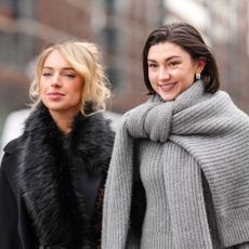 A guest wears black oversized Michael Kors coat, black faux fur scarf, black sheer mesh floral lace midi Michael Kors skirt, shiny black Michael Kors leather bag. Kate Bartlett wears black sunglasses, silver earrings, dark gray oversized knit Michael Kors sweater, light gray long sleeve Michael Kors sweater, dark gray midi Michael Kors skirt, white leather Michael Kors bag with silver chains, outside Michael Kors, during the New York Fashion week Fall/Winter 2025 on February 11, 2025 in New York, New York