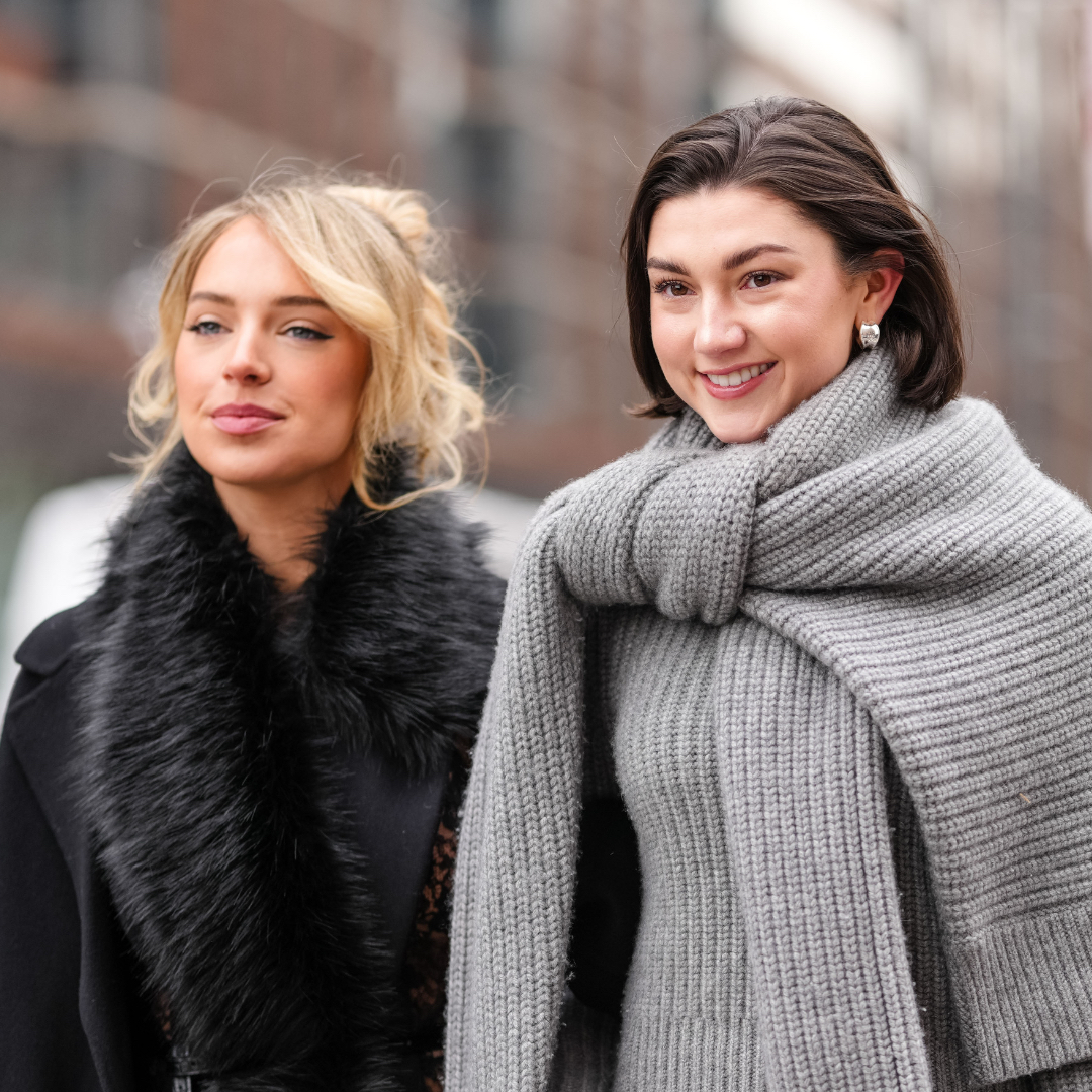 A guest wears black oversized Michael Kors coat, black faux fur scarf, black sheer mesh floral lace midi Michael Kors skirt, shiny black Michael Kors leather bag. Kate Bartlett wears black sunglasses, silver earrings, dark gray oversized knit Michael Kors sweater, light gray long sleeve Michael Kors sweater, dark gray midi Michael Kors skirt, white leather Michael Kors bag with silver chains, outside Michael Kors, during the New York Fashion week Fall/Winter 2025 on February 11, 2025 in New York, New York