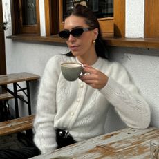 British style influencer Jessica Skye sits at an outdoor cafe drinking a cup of coffee wearing black oval sunglasses, a fuzz off white cardigan, studded belt and leather pants