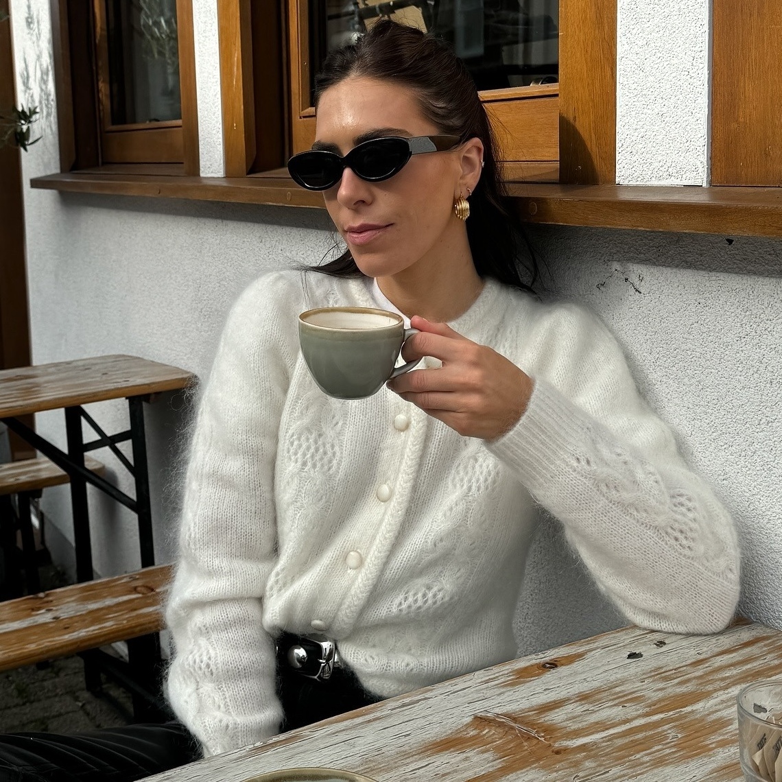 British style influencer Jessica Skye sits at an outdoor cafe drinking a cup of coffee wearing black oval sunglasses, a fuzz off white cardigan, studded belt and leather pants