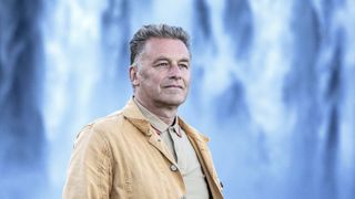 Chris Packham in a mustard jacket stands in front of a waterfall in Earth.