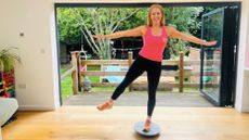 Woman balances on one leg on a balance board. She is smiling and wearing a pink tank top and black leggings.