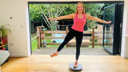 Woman balances on one leg on a balance board. She is smiling and wearing a pink tank top and black leggings.