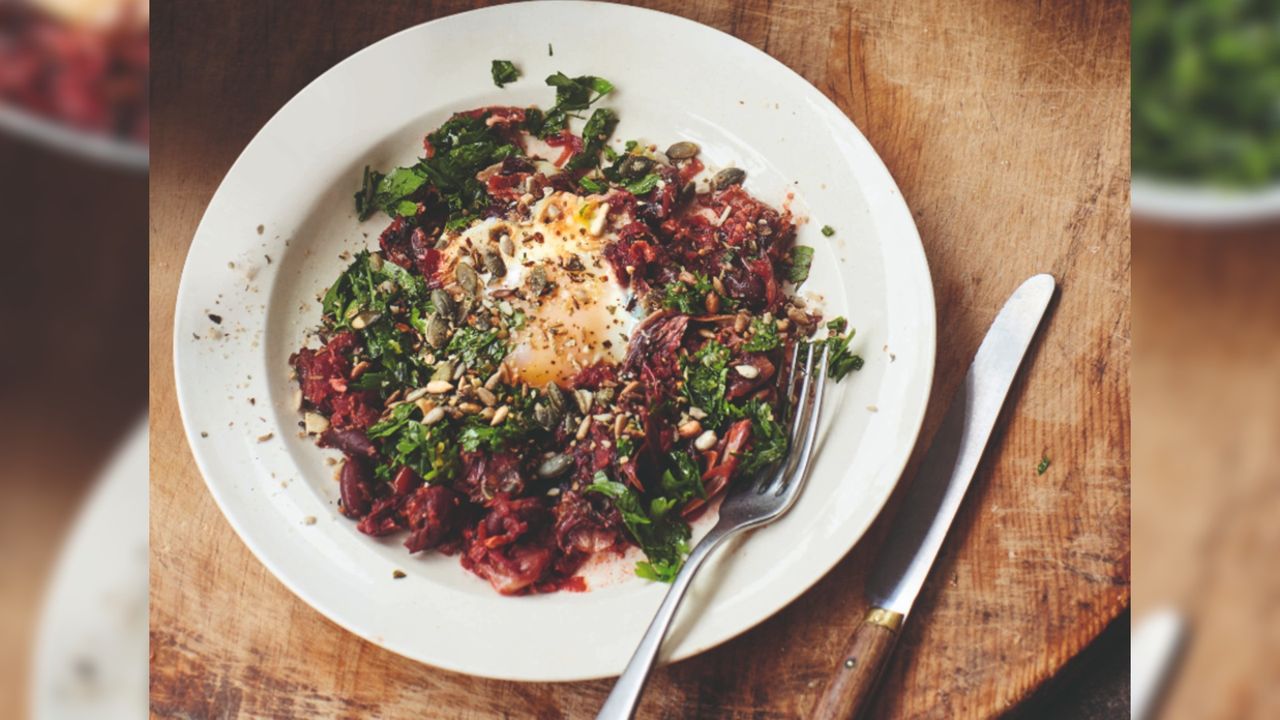 Photo of purple shakshuka on a plate. There is a fork on the plate and knife next to the plate on a wooden table