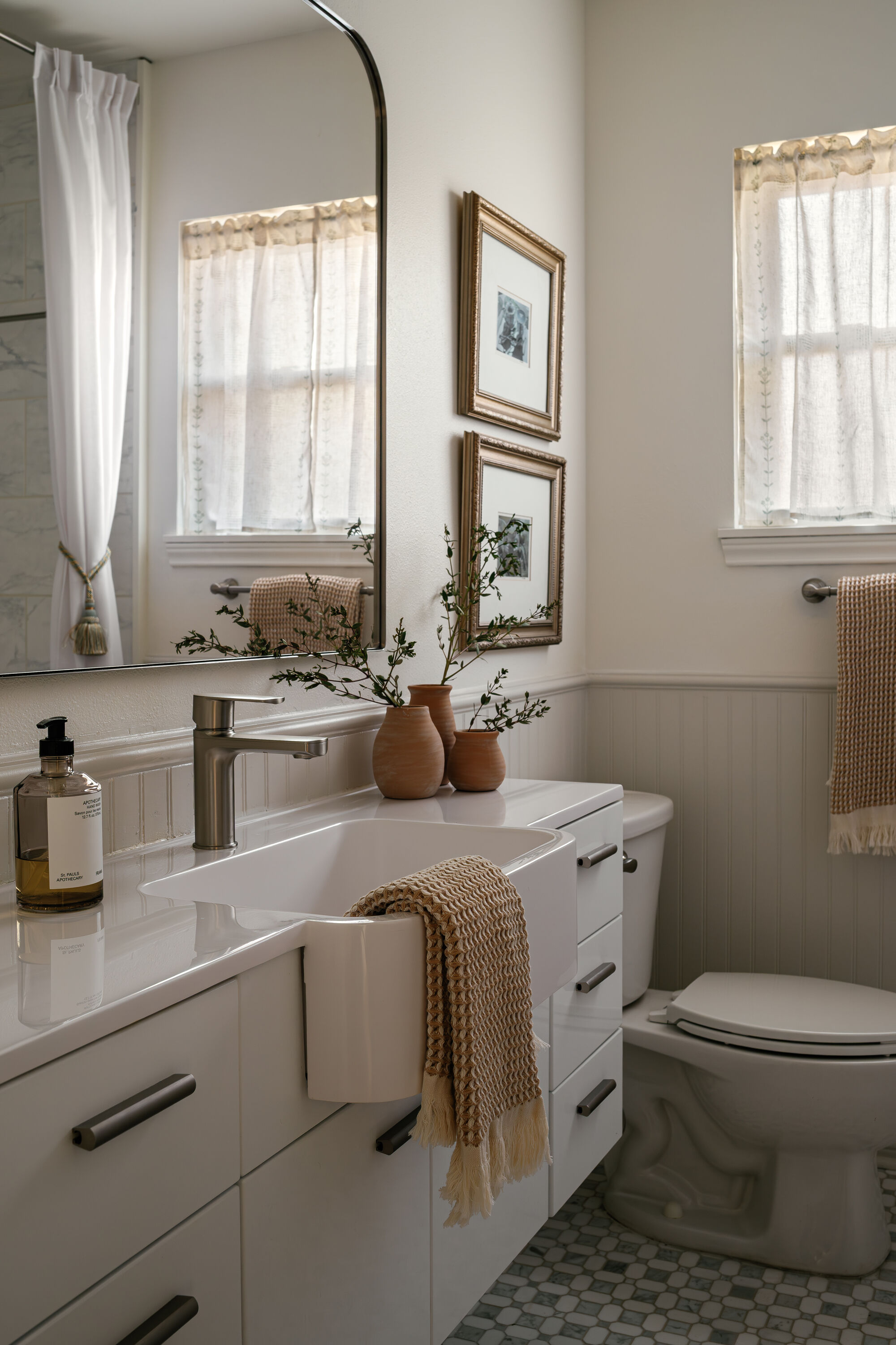 A neutral bathroom with a tub sink that have a texted, caramel hand towel hung over the edge. There are three small terracotta vases on the counter and two art pieces hanging above the toilet.