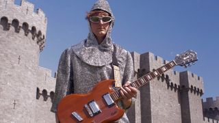 Will Paquin wears a chain mail suit and sunglasses and holds a Guild Polara in Cherry Red in front of a castle.