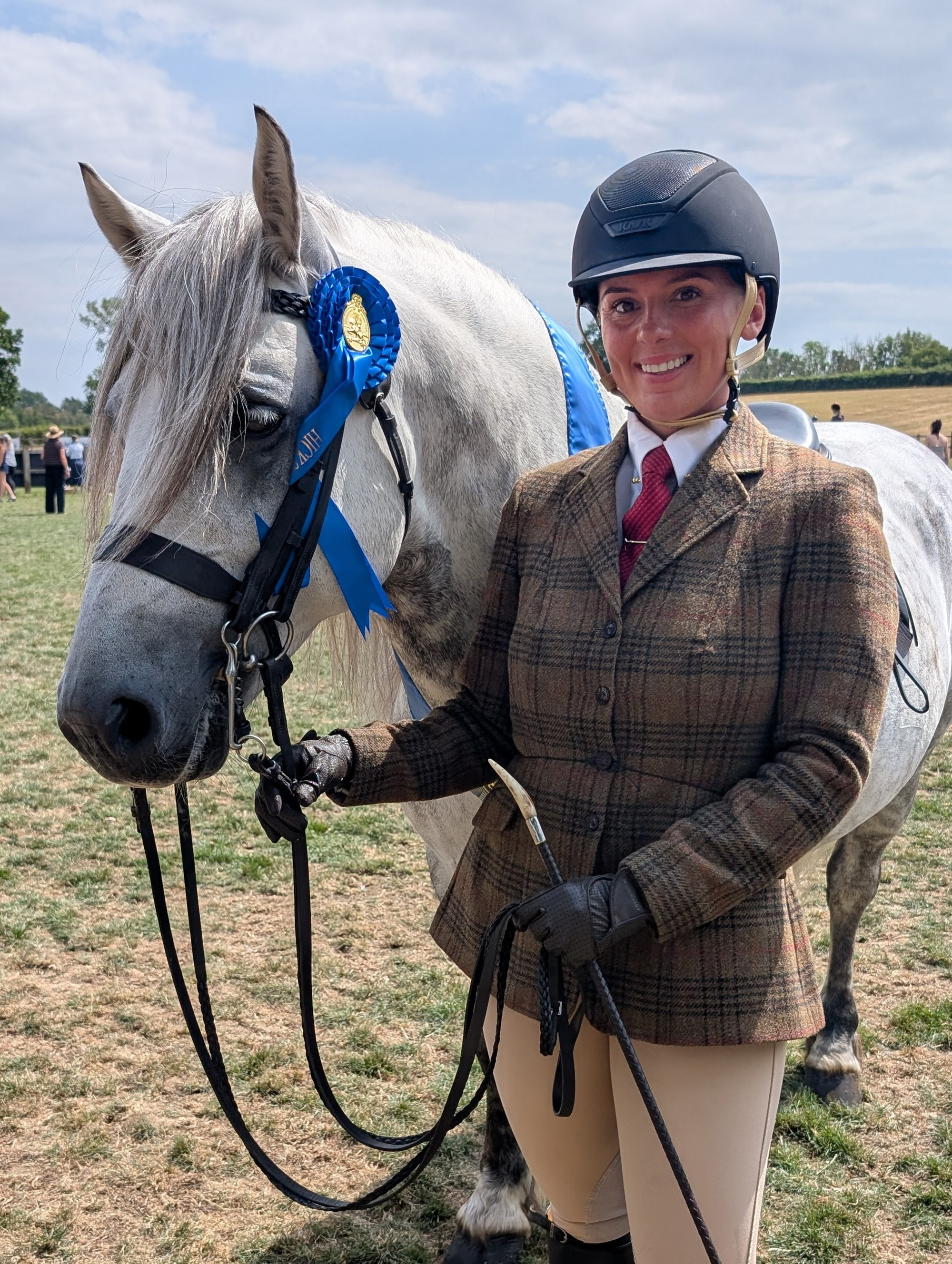 A grey Highland pony wearing his blue winners rosette and sash standing with her rider wearing tweed.