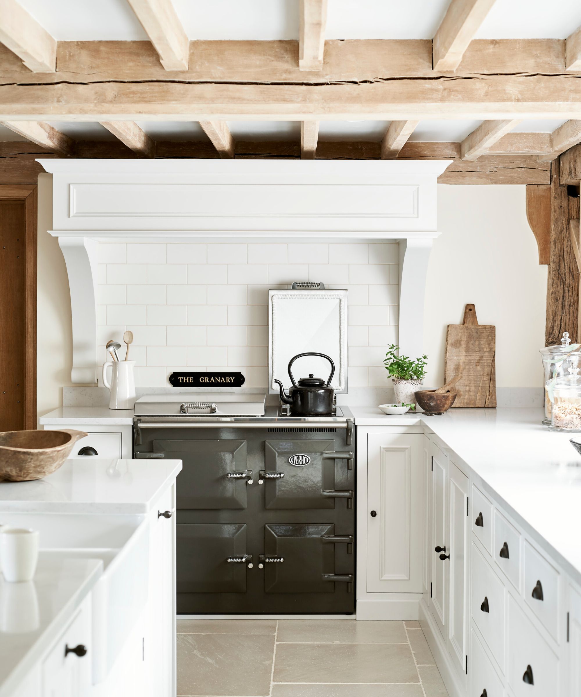 A traditional kitchen features a dark grey range cooker framed by white cabinetry and subway tiles. A black kettle rests on the hob. Wooden boards, ceramics, and simple greenery add texture, creating a calm, pared-back space with classic farmhouse character.