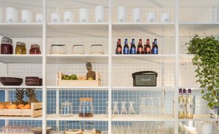 View of the white metal shelves in the Deli restaurant. They're filled with various fruits in baskets, glasses, jars, plates, and alcohol bottles.