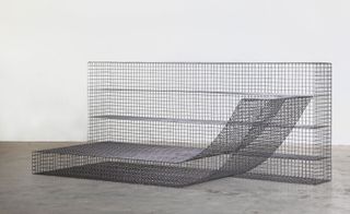 Grey modular lounge seating based on a grid structure, made of metallic net photographed against a wall like structure made of metallic net in a room with white walls and grey floor.