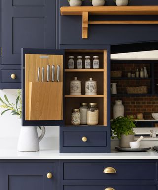 A kitchen with navy blue kitchen cabinets on the wall and below the white countertop, with the one in the middle open revealing a light wooden door and three shelves with jars on them, with further kitchen space beyond