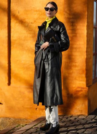 A guest wears black sunglasses, gold earrings, a yellow turtleneck pullover, a black shiny leather belted long coat, a black shiny leather handbag from Loewe, black and white shiny leather pointed / block heels knees boots , outside Mark Kenly Domino Tan, during the Copenhagen Fashion Week Autumn/Winter 2023 on February 02, 2023 in Copenhagen, Denmark.