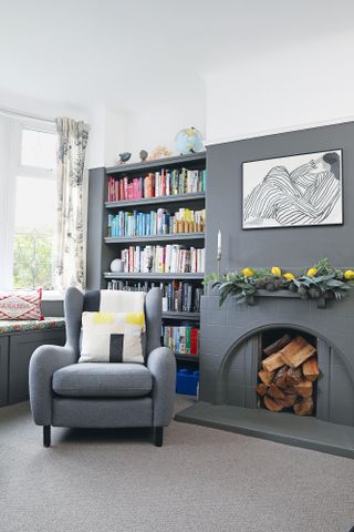 Grey living room with open shelving full of books, grey armchair, and fireplace filled with logs, with framed monochrome line drawing above