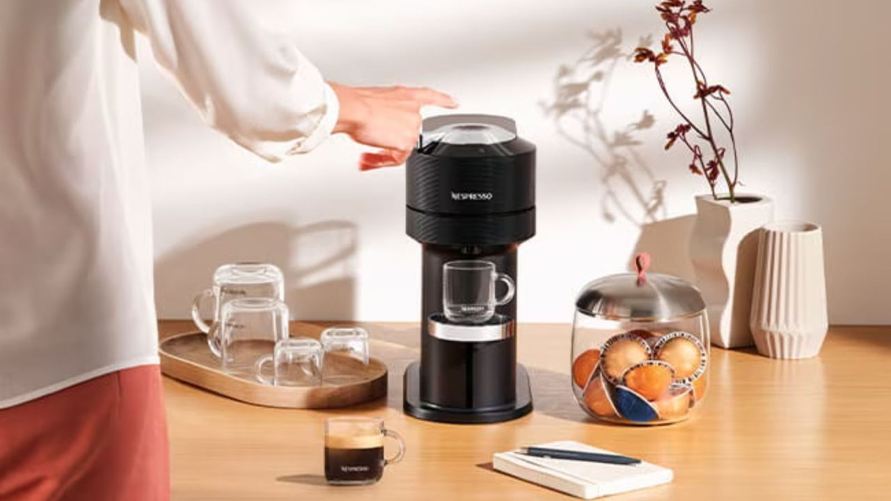 A woman in a white shirt and pink trousers pressing a button on a Nespresso coffee maker on a wooden work surface, surrounded by pots of pods and glass mugs. 