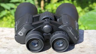 EclipSmart 12x50 binoculars placed on a wooden surface with green foliage behind