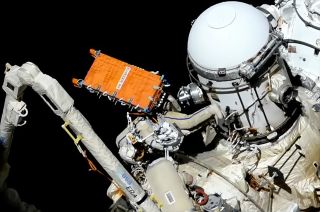 an astronaut in a white spacesuit installs an orange radar communications system on the international space station.