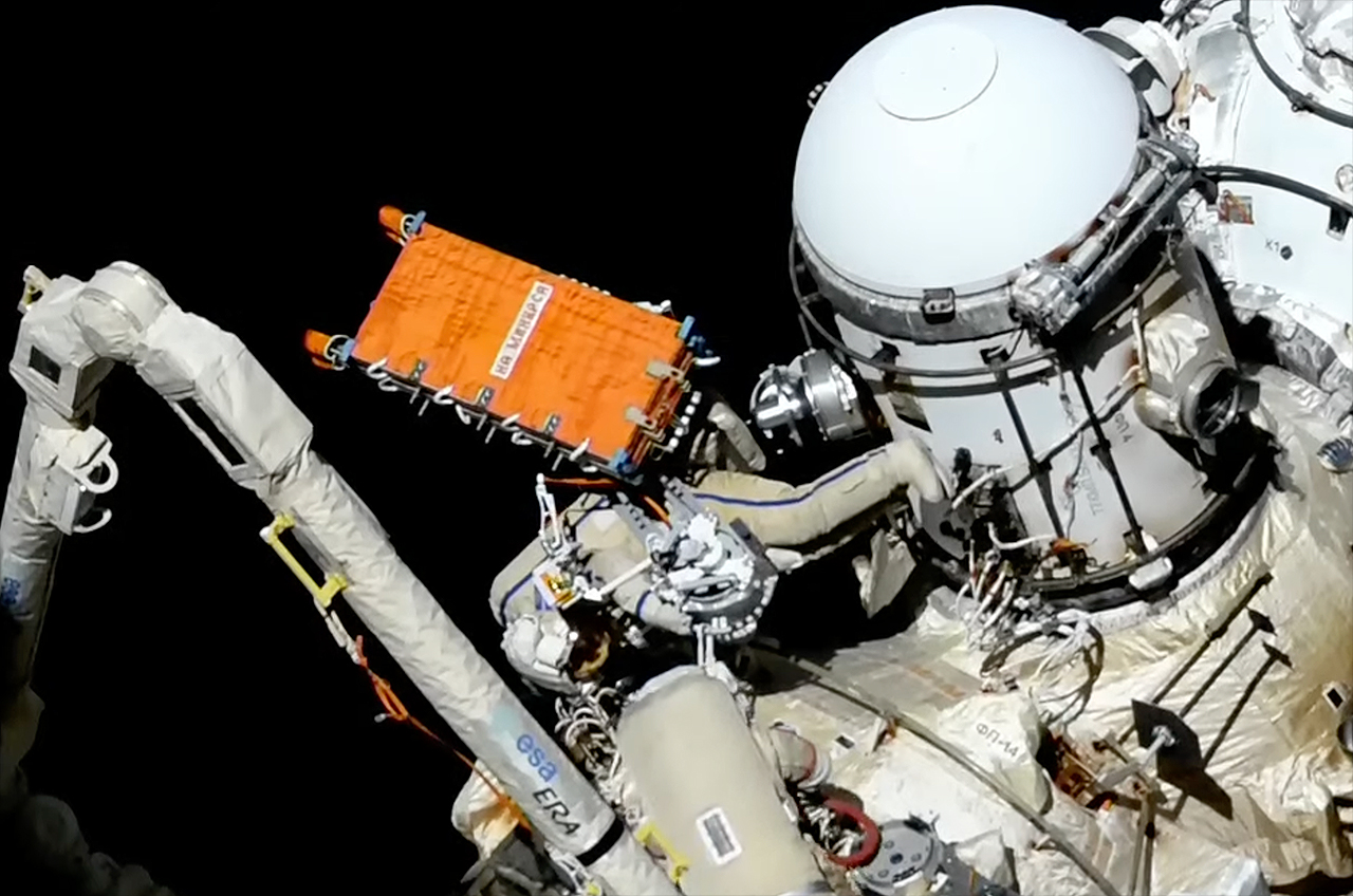 Seorang astronot yang mengenakan pakaian antariksa putih memasang sistem komunikasi radar oranye di Stasiun Luar Angkasa Internasional.