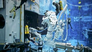 A person wearing a white space suit floats underwater with divers nearby interacting with various equipment