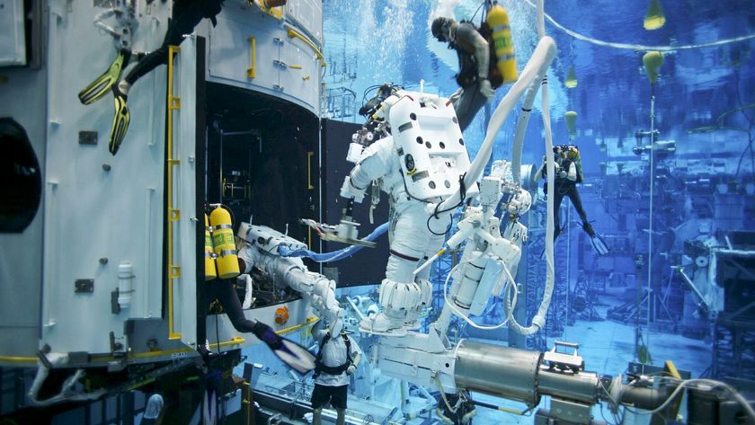 A person wearing a white space suit floats underwater with divers nearby interacting with various equipment