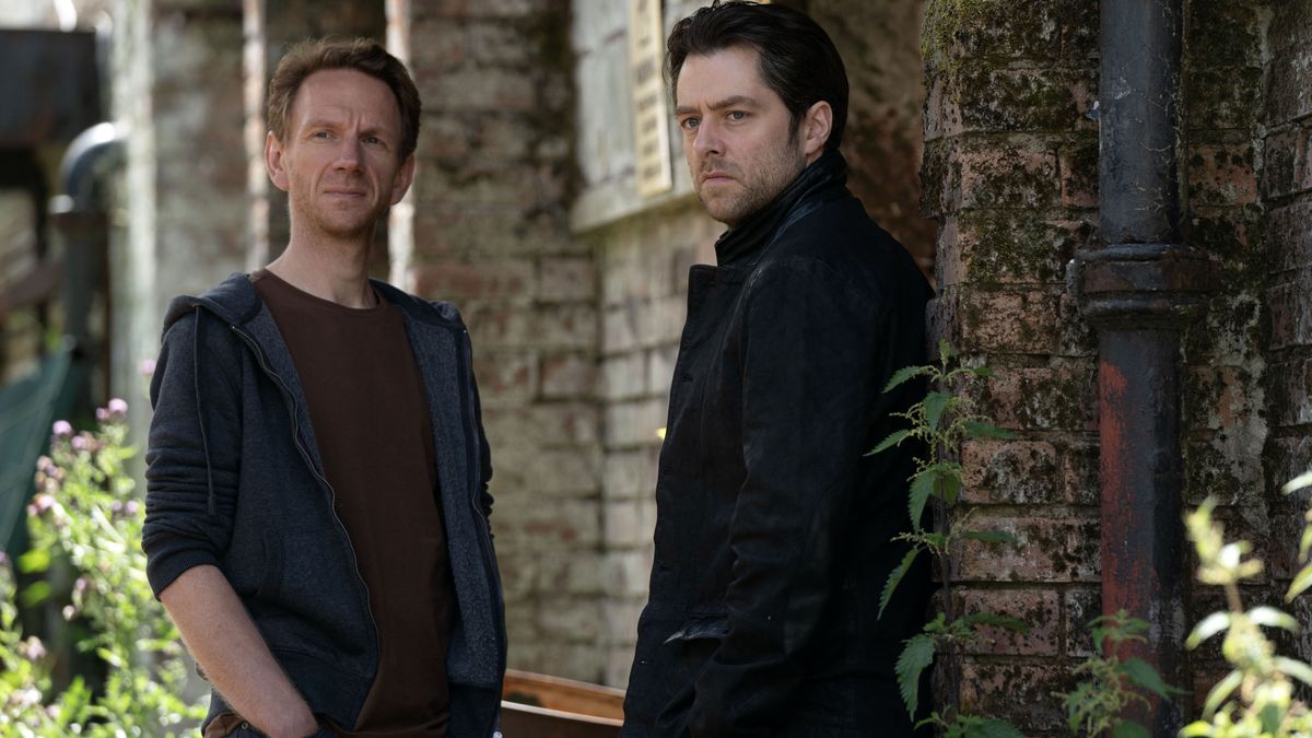 Brian Ferguson as Michael Rebus and Richard Rankin as John Rebus stand by a wall in Rebus.