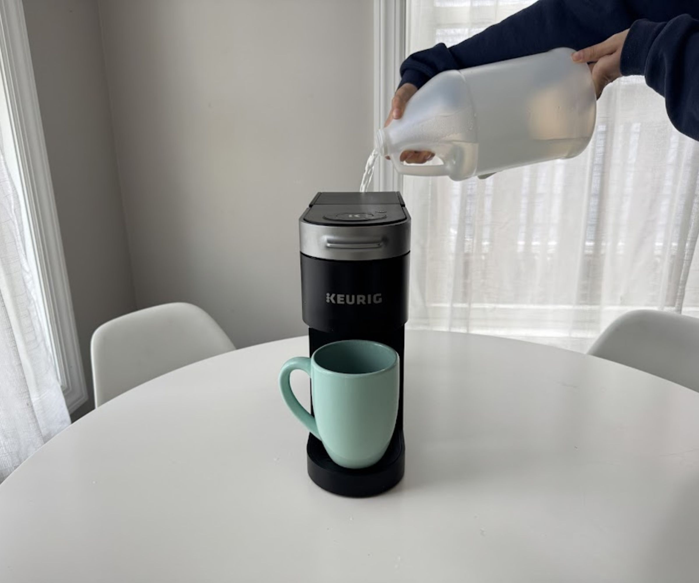 Someone pouring a clear liquid from a milky-white plastic bottle into the reservoir of a black Keurig coffee machine, with a blue cup under the spout.