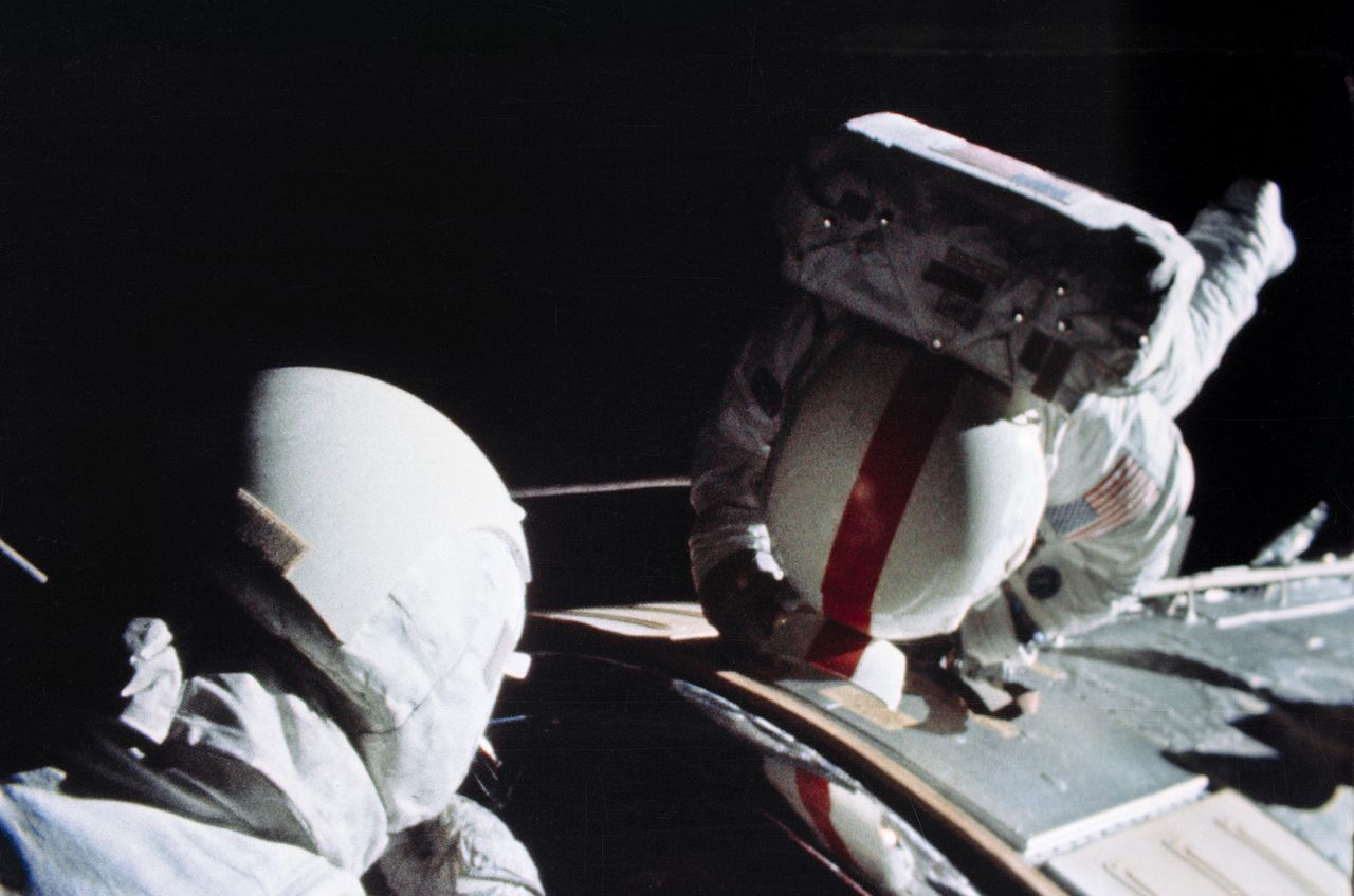 two astronauts in white spacesuits work outside a capsule during a spacewalk.