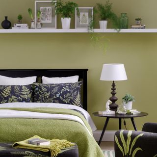 Green nature patterned bedroom, fitted shelf, black botanical armchair and matching footstool and pillows, round dark wood bedside table.