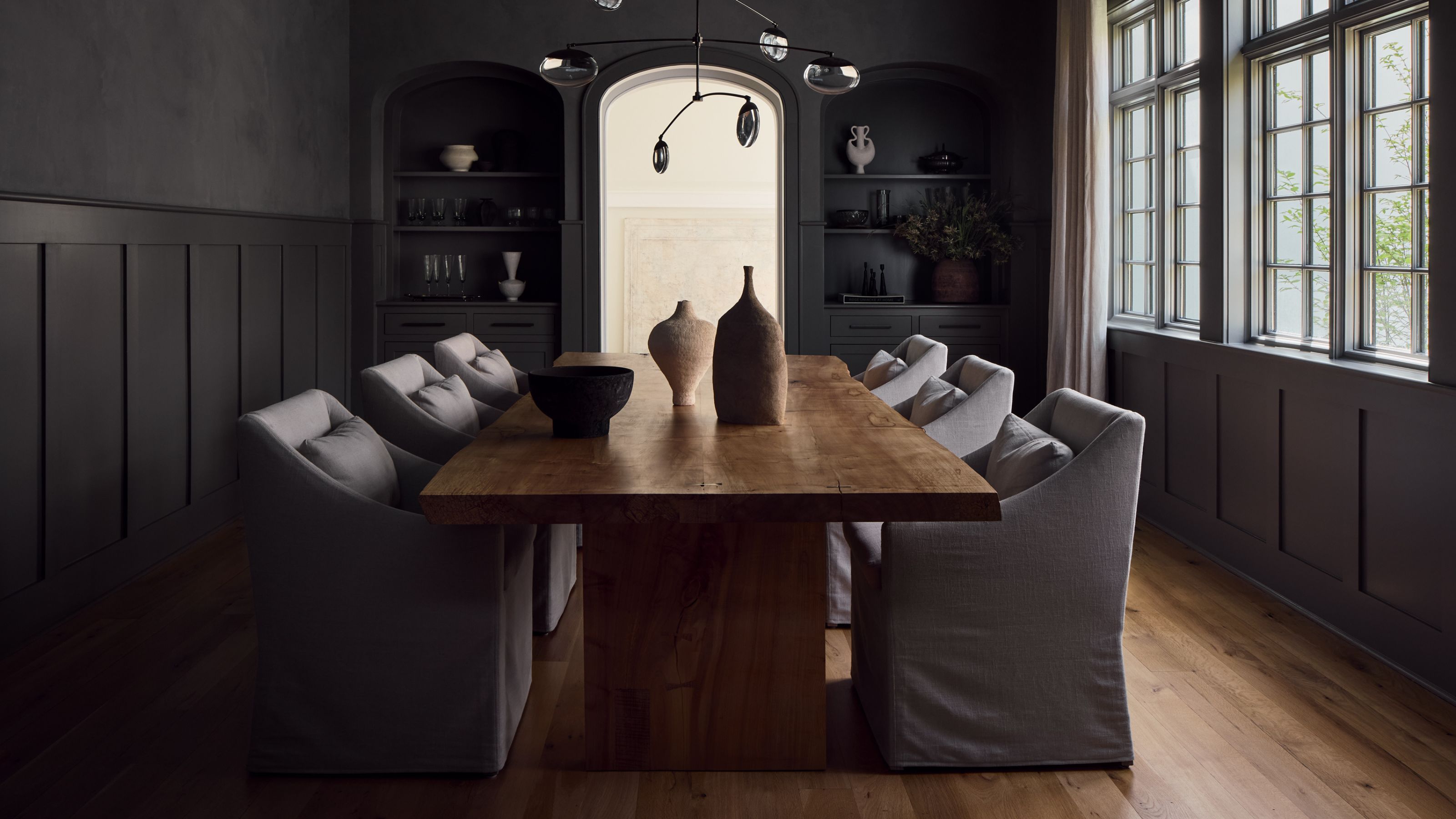 Dark dining room painted in a moody charcoal gray with the same colored paneling half way up the walls. There&#039;s a large wooden table in the middle with gray upholstered dining room chairs in the same gray and a large window fills the room with light