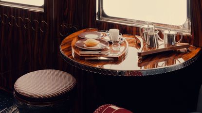A window seat in a luxury train carriage, featuring sleek breakfast tableware, lacquered marquetry, and upholstered stools with checkered motifs in red and blue.