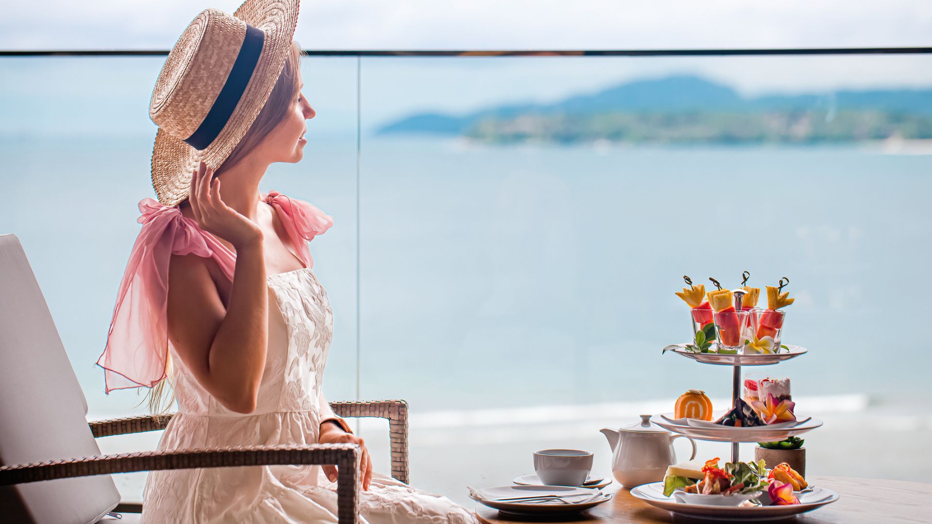 A woman sits in front of a sea view with an afternoon tea