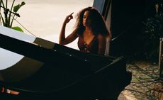 View of Solange sitting at a black piano in a space with a large curved window and green plants