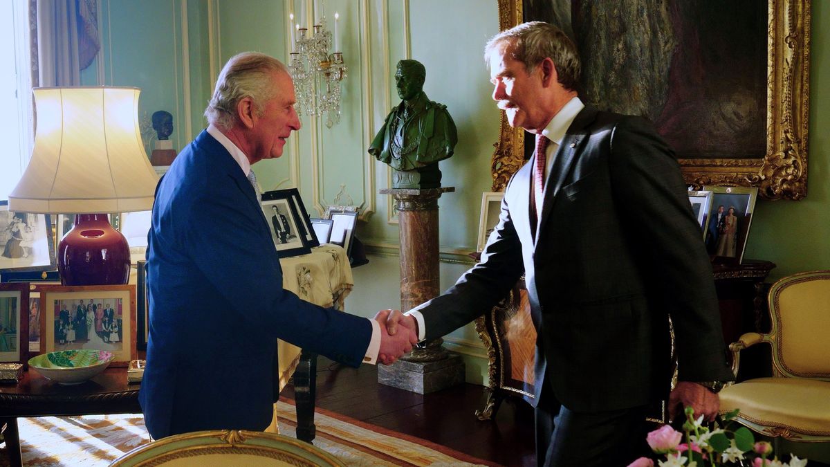 king charles iii shaking hands with chris hadfield in a palace room surrounded by photos, chairs, tables. a statue is on a pillar between them